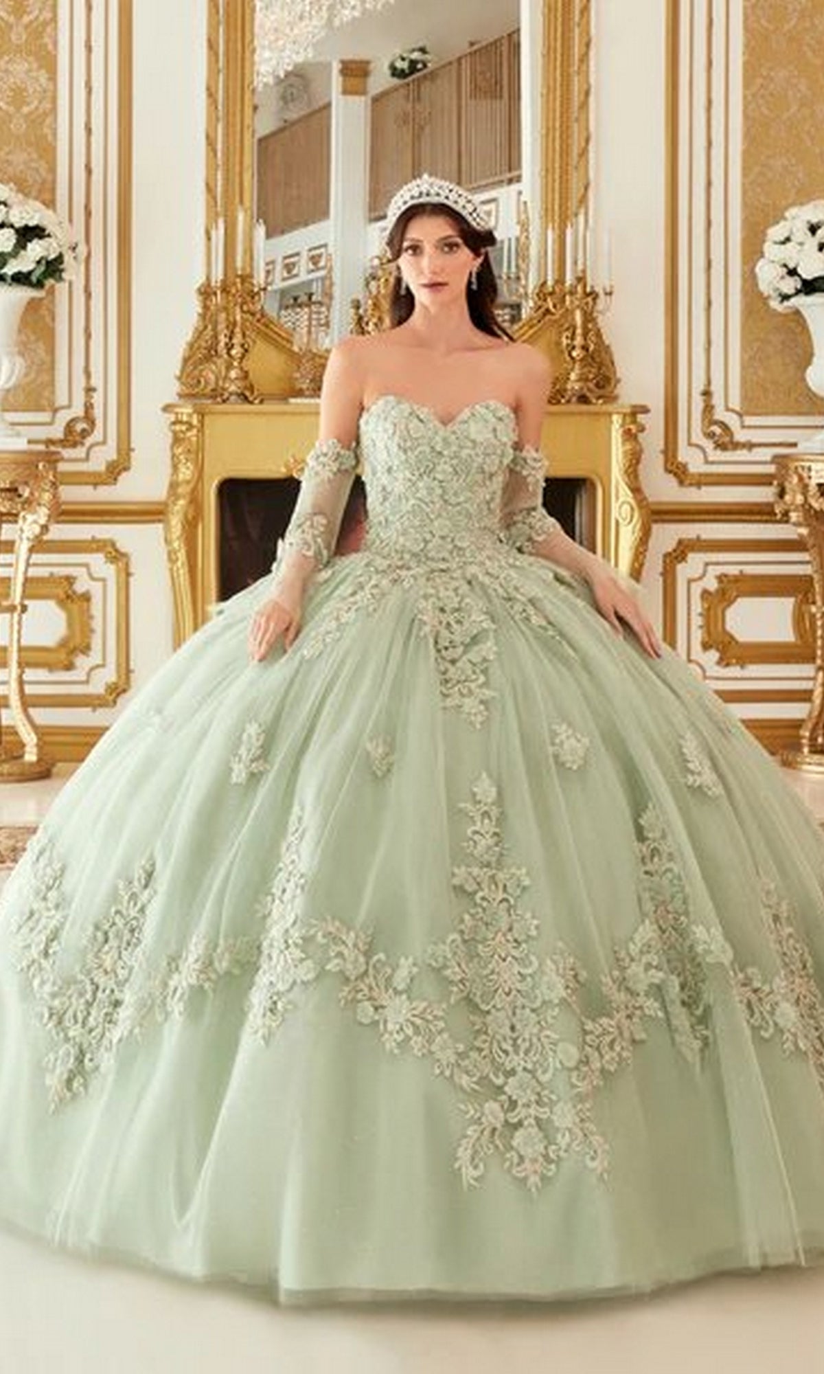 A woman wearing the Quinceanera Dress 15714 by Ladivine, a pale green pastel ball gown with delicate lace embroidery and a tiara, stands in an opulent, gold-decorated room.